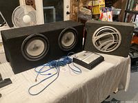 Two subwoofer speaker boxes on table with amplifier and blue wires. JL Audio subwoofers and Q Logic visible.