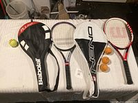Lot of 3 tennis rackets displayed on a surface with three racket covers and four tennis balls.