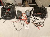 Wide view showing three electrical tools: digital multimeter with red and black leads, battery charger with clamps and display, and a handheld inflator device with hose.