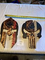Two handmade wooden wall art pieces including a Spartan helmet and a skull, both with maple leaf carvings, on white fabric background next to a measuring tape showing sizes.
