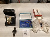 Photo of lot with gold-tone wristwatch in box, silver-tone wristwatch, Barbie Keepsake ornament, and keychain in blue box on bed.