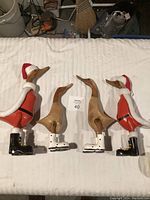 Four wooden duck figurines arranged on a white surface; two larger ducks in Santa outfits and two smaller natural wood ducks with white boots with red dots.