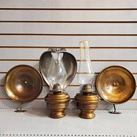 Front view of both brass wall-mount oil lamps, chimneys, soot catchers, and reflector arranged on shelf