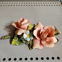 Close-up photo of the porcelain flower arrangement showing two pink roses, white flowers with yellow centers, and green leaves with some chips.