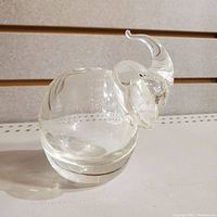 Clear glass elephant figurine with prominent ears, raised trunk, and round body, shown from side angle on white surface.