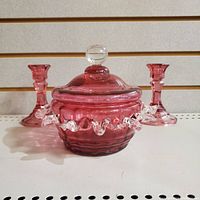 Front view of lidded cranberry glass dish with two cranberry glass candleholders behind, showing color and applied clear ruffle detail