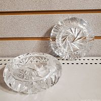 Photo showing two clear crystal ash trays, one upright and one lying flat, both with pinwheel pattern and visible facets.