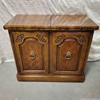 Front view of closed wooden bar cabinet with decorative panel doors, metal ring pull handles, and scalloped top edge.