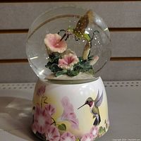 Snow globe featuring a hummingbird inside with flowers, clear glass globe sitting on a painted base with floral and hummingbird design