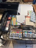 Overall art supplies lot including toolbox, brushes, colored pencils, small canvases, and paint tubes laid out on a surface
