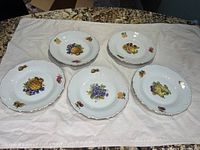 Five vintage Czechoslovakia fruit plates laid out on white cloth showing front surfaces with fruit motifs and gold trimmed scalloped edges.