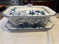 Side view of rectangular casserole with lid on platter showing blue floral and fruit designs, raised lid knob, and blue rim highlights
