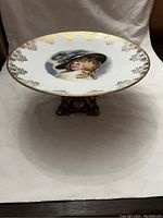 Front view of round Limoges porcelain cake plate with hand-painted portrait and gold leaf embellishments on rim mounted on ornate bronze pedestal base.