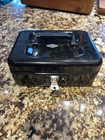 Black metal locking cash box with a handle and lock, closed and resting on a granite countertop.