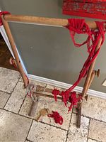 Wooden Ram Loom with red yarn stretched across the frame, viewed from an angle showing the full loom structure on a tiled floor.