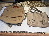 Two bags laid out on a white cloth against a granite surface: a beige canvas messenger bag/backpack with front flap and external zippered pockets on the left, and a vintage light tan leather briefcase handbag with buckled straps on the right.