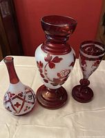Three hand blown glass pieces including a white and red rounded vase, a goblet, and a decorative bottle.