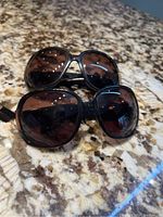 Two pairs of black-framed dark-lensed sunglasses on a granite surface with one pair closer to the camera and some light reflections visible.