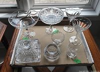 Eight crystal pieces on a cardboard surface by window with natural light showing footed bowls, stemware, covered dish, candle holder and bud vase.