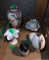 All five items displayed in a cardboard tray on a wooden surface against a concrete wall; includes the leaf dish, milk glass bud vase, ceramic sculpture, Asian vase, and small lidded jar