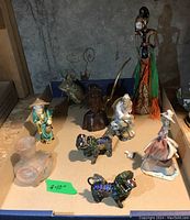 Full view of the eight figures arranged on cardboard showing size range and diversity of items including foo dogs, fisherman, wooden bust, bud vase, cat, and two female figures including a tall colorful wire figure.