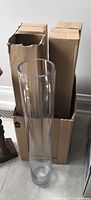 Tall clear glass floor vase out of box standing upright in front of two cardboard boxes containing sealed vases.