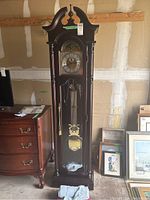 Front view of the grandfather clock showing cherry wood case, brass face, pendulum, and key inside glass front panel.