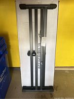 Photo showing full metal keyboard stand folded and leaning against a white box with yellow wall background. New and still in original packaging.