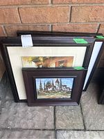 Five framed landscape prints leaning against a brick wall, showing different sizes of frames with dark woods and matting for some.