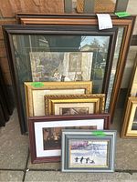 Six framed prints of varying sizes stacked together outside on pavement. Frames include dark wood, brown, gold, and gray colors.