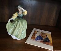 Porcelain figurine Buttercup posed on wooden shelf next to Royal Doulton Pretty Ladies catalog