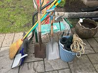 Yard and garden tools including two shovels, broom, dustpan, blue bucket with smaller pots, and extension cords.