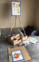 Full view of wooden tripod easel with clipped watercolor painting, wooden travel art case with open drawers showing some supplies, additional wooden storage box, clipboard with another painting taped, and loose papers on floor.
