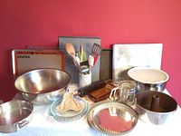 Full lot of various baking supplies pictured together showing metal bowls, baking pans, cookie sheets, utensils, and a sifter against a red wall.