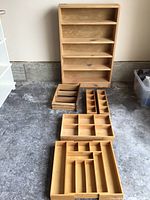 Full view of the wooden bookcase with five fixed shelves next to five bamboo drawer organizers of different compartment layouts arranged on concrete floor.