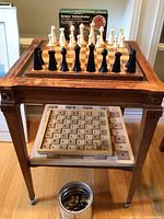 Full view of vintage game table with two wooden pegged chess boards on lower shelf, largest chess board on table top with black and white chess pieces, metal container with pieces on floor beneath.