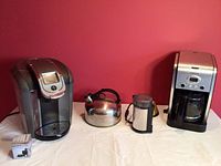Wide shot showing Keurig 2.0 brewer, stainless steel teapot, Krups grinder, and Cuisinart coffee maker along with box of filters on white tablecloth against red background.