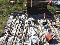 Overhead view of multiple long-handled gardening tools laid on tarp, with wooden garden cart visible