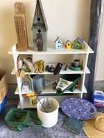 Full view of white shelf with bird houses on top, gardening gloves, hand tools, watering cans, and a small wheelbarrow on shelf and floor.