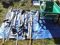 Full collection of long-handled tools laid on blue tarp next to cart