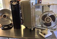 Front view showing all 5 appliances on table: HeatDish heater, ThermaPure air purifier, white oil radiator heater, white box fan, and white oscillating desk fan.
