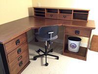 Full view of L-shaped corner desk and black office chair. Desk in medium hazelnut brown veneer with three drawers on left side, cabinet on right side, and removable hutch on top.