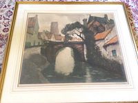Framed lithograph showing a European town scene with a stone bridge and buildings with orange roofs over water.