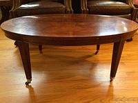 Oval wood coffee table with wood grain top, visible legs with caster wheels, and leather chairs in background.