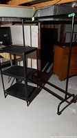 Clothes rack and metal storage shelf side by side on a basement floor. Clothes rack has horizontal bar and wheels. Shelf unit has three shelves.