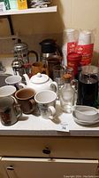 Overview of all coffee and tea related items on kitchen counter including mugs, coffee presses, grinder and glassware.