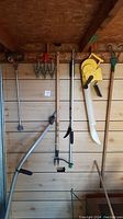 Full view of gardening tools hanging on a wooden wall inside a shed, showing aerator, seed spreader, hoe, sickle, and cultivator