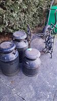 Photo showing three metal milk jugs with weathered surfaces next to a green wheelbarrow.