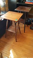Two wooden pedestal/plant stand tables side by side showing bobbin legs, square tops, and lower shelves on hardwood floor.