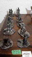 View of twelve pewter miniature figurines arranged in two rows on wooden surface, all with bases and detailed poses, some sitting, standing, or pulling equipment
